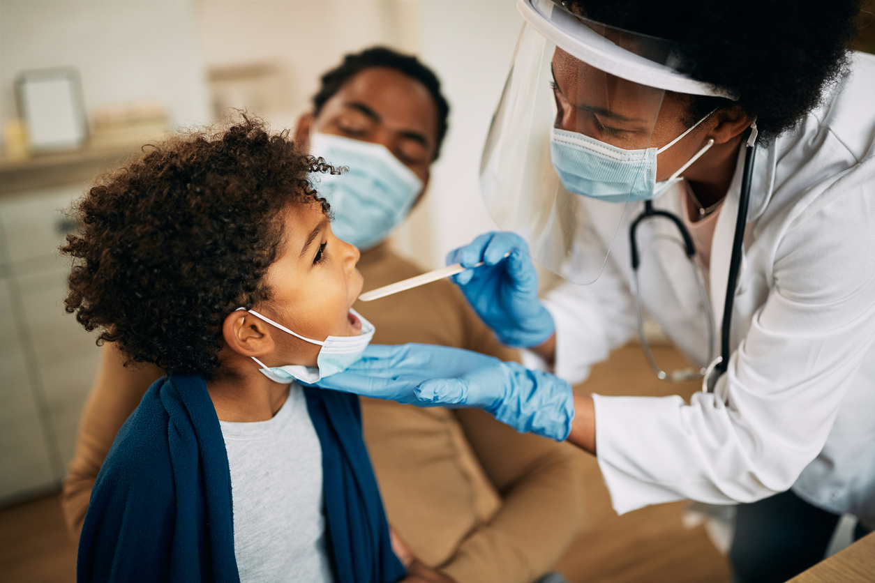 A female doctor wearing gloves, a face shield, and a mask performs a preventative care checkup on a child. 
